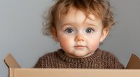 Adorable toddler with bright blue eyes and curly hair wearing brown knit sweater peeks out from cardboard box, expressing curiosity and innocence against gray background.