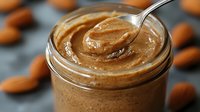 Creamy almond butter in glass jar with spoon showing smooth texture and whole almonds scattered around on dark background, macro food photography.