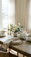 Elegant dining table setting with white roses centerpiece, neutral tableware, linen napkins and wine glasses in bright natural light interior.