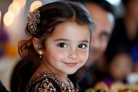 Young girl with dark hair wearing elegant brown lace dress and ornate hair accessory smiles warmly at special occasion celebration with soft bokeh background.