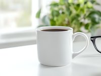 White ceramic mug filled with dark coffee on bright windowsill with green plants and reading glasses, creating cozy morning atmosphere.