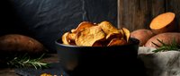 Crispy golden potato chips in rustic black bowl with fresh herbs and whole potatoes on dark wooden background for snack food and cooking concepts.
