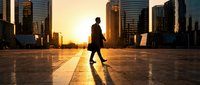Silhouette of a male professional in business attire walking across an empty urban plaza during golden hour sunrise, with modern skyscrapers in background.
