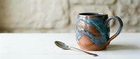 Handcrafted ceramic mug with blue and orange glazed finish sits beside copper spoon on white surface against textured stone wall background.