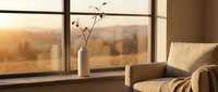 Cozy living room interior with armchair and vase by large window overlooking autumn landscape at golden hour sunset, creating warm peaceful home atmosphere.
