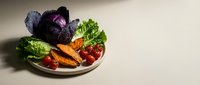 Fresh organic vegetables arranged on ceramic plate including purple cabbage, leafy greens, carrots, and cherry tomatoes for healthy cooking and nutrition.
