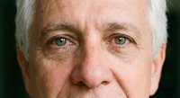 Close-up portrait of mature Caucasian man with gray hair and green eyes showing natural aging lines and wrinkles on face against dark background.