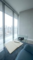 Modern minimalist home office with glass desk, open book and headphones overlooking urban cityscape through floor-to-ceiling windows for remote work concepts.