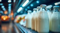Glass milk bottles with metal caps lined up on refrigerated supermarket shelf with bokeh lights in background.