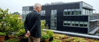 Senior man with white hair, Caucasian ethnicity, wearing dark business casual attire, observing modern sustainable architecture with green roof garden and black facade building.