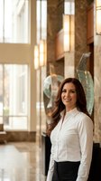 Professional businesswoman with long brown hair smiling confidently in modern office lobby with warm lighting and contemporary architecture.
