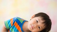 Young child with dark hair and light skin tone wearing colorful striped shirt, lying down against soft pastel background. Suitable for parenting, childcare, education, and family-related marketing materials.