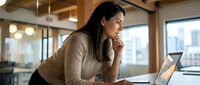 Woman of South Asian descent, professional appearance, sitting at desk in modern office with laptop, hand to face in thoughtful or concerned expression, wearing beige sweater, natural lighting from windows, urban cityscape visible in background, contemporary workplace setting.