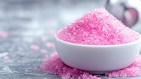 Pink aromatic bath salt crystals in white ceramic bowl on gray stone background with scattered grains, spa and wellness concept for beauty treatments.