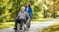 Senior Caucasian man in wheelchair with caring middle-aged woman caregiver walking together in sunny park pathway surrounded by green trees and foliage.