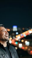 Middle-aged man with dark hair and beard, wearing black leather jacket, smiling upward at night with blurred city lights in background. Use for lifestyle, optimism, urban themes.
