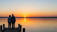 Couple standing on wooden dock watching golden sunset over calm lake water creating romantic peaceful moment together during evening twilight hours.