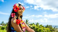 Young woman with long dark hair and warm complexion wearing vibrant red and yellow flowers in hair and lei garland, smiling outdoors with tropical landscape and blue sky background for travel, culture, and lifestyle content.