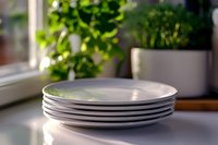 Stack of clean white ceramic plates on kitchen counter near window with green plants, soft natural daylight creates gentle shadows and bokeh effect.