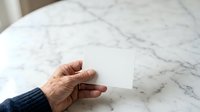 Mature adult hand holding blank white card on marble surface, suitable for business mockups, greeting cards, invitations, and promotional designs.