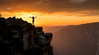Person celebrating success on mountain cliff edge during golden sunset with arms raised in triumph over dramatic canyon landscape for motivation concepts.