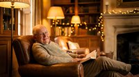 Senior man reading book in cozy living room with warm lighting and fireplace during evening relaxation time at home.