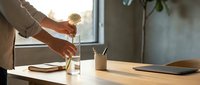 Professional woman pouring fresh coffee from glass pot into white mug at modern office desk during morning routine creating productive workspace atmosphere.