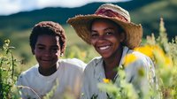 Young African American woman wearing straw hat and light blue shirt smiling in wildflower field with young boy in white shirt, mountains in background, outdoor summer portrait.