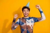 Happy young Caucasian man in party hat celebrating with raised fists against bright orange background. Joyful birthday celebration concept.