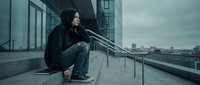 Young man in black hoodie sitting alone on urban rooftop terrace overlooking city skyline expressing loneliness and contemplation in moody atmosphere.