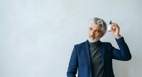 Mature man with gray hair and beard, wearing navy blazer and dark turtleneck, adjusting glasses against white background. Suitable for business, corporate, professional, and lifestyle design projects.