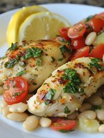 Grilled chicken breast fillets with herb seasoning served with white beans, fresh tomato slices, cucumber, and lemon wedges on white plate.