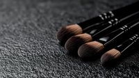 Professional makeup brushes with soft brown bristles arranged in row on dark textured background, dramatic lighting highlights quality cosmetic tools.