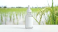 Frosted glass dropper bottle with blank label on white surface, green rice field background, natural skincare product mockup.