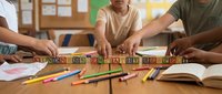 Children hands arranging wooden alphabet blocks spelling BUSINESS on classroom desk with colorful pencils and notebooks around.