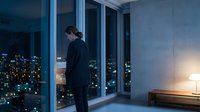 Young businessman in dark suit standing by floor-to-ceiling windows overlooking illuminated city skyline at night in modern office or apartment interior.