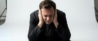 Stressed businessman in dark suit holding head in hands showing mental exhaustion and workplace pressure in modern office environment.