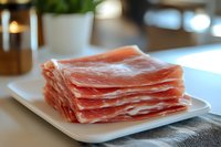 Fresh sliced prosciutto ham arranged on white plate in modern kitchen setting for gourmet cooking and culinary presentations.