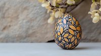 Decorative Easter egg painted with blue floral pattern on orange background, displayed with white spring blossoms against textured wall. Seasonal holiday composition.