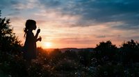 Young girl with curly hair blowing bubbles in meadow at golden sunset, silhouetted against warm sky with trees and hills, capturing childhood wonder and outdoor leisure moments.