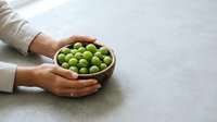 Hands holding wooden bowl filled with fresh green apples on gray concrete surface, suitable for food, agriculture, harvest, and organic produce marketing materials.
