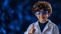 Young Hispanic boy wearing lab coat and safety glasses shows thumbs up against blue bokeh background, expressing confidence in science education.