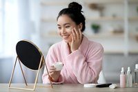 Happy Asian woman applying skincare cream while looking in mirror during morning beauty routine at home vanity table with cosmetic products.