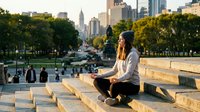 Young woman sitting on stone steps in urban park during golden hour sunset with city skyline and skyscrapers in background, peaceful moment in metropolitan setting.