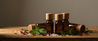 Three amber glass essential oil bottles with wooden caps on rustic table, surrounded by mint leaves, dried petals, and sea salt crystals.