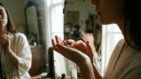 Woman with long dark hair and light skin tone examining her hands in mirror, wearing beige clothing in bright bathroom setting with natural window light, reflecting on skincare routine.