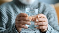Person in gray knit sweater holding clear glass of sparkling water with bubbles, close-up view of hands. Soft natural lighting creates cozy atmosphere for health and wellness content.