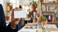 Person with dark skin tone holding blank white business card in modern office workspace with bookshelves and plants, suitable for corporate branding and professional networking materials.