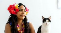 Young woman with dark long hair and medium skin tone wearing vibrant red and yellow flowers in her hair and floral lei, posing alongside black and white cat against white background.