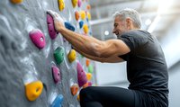 Mature athletic Caucasian male with gray hair climbing indoor rock wall, wearing black sportswear, focused on colorful climbing holds in modern gym facility.